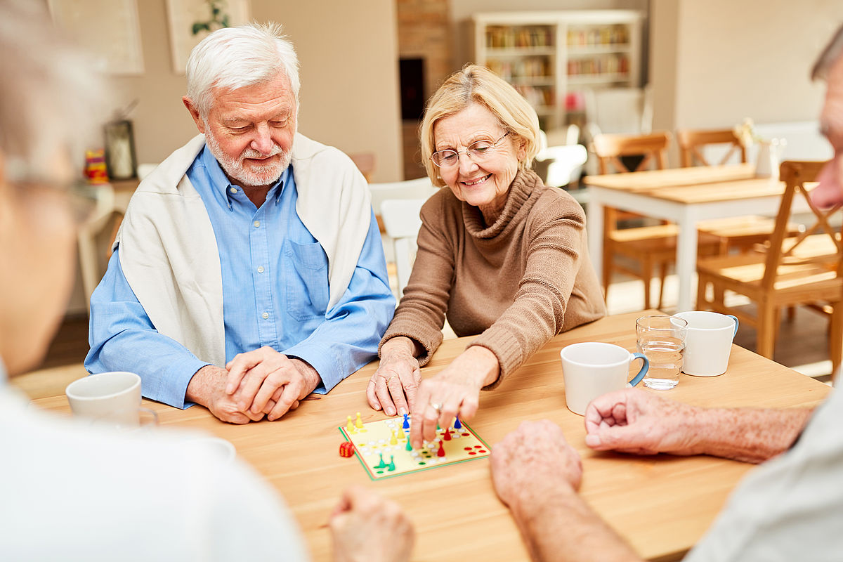 Vier Senioren an einem Holztisch, die miteinander das Brettspiel "Mensch-ärgere-Dich-nicht" spielen. Im Hintergrund sind scharf ein bärtiger weißhaariger Mann in blauem Hemd und übergehängten beigefarbenem Pullover zu sehen, der mit aufeinandergelegten Händen am Tisch sitzt und auf das Spielbrett blickt. Neben ihm sitzt eine Senioren mit blondem Haar und Brille, die einen braunen Strick-Rollkragenpullover trägt. Sie lächelt und bewegt mit der linken Hand eine spielfigur auf dem Spielbrett. Im Vordergrund sind im Anschnitt zwei weitere Personen am Tisch sitzend erkennbar, offensichtlich ebenfalls Senioren. Im Hintergrund ist eine Esstischgruppe, ein Sideboard mit Büchern und Bilder an der hellen Wand zu sehen.