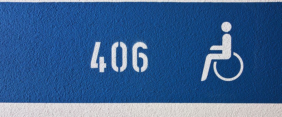 Blaue Parkhausmarkierung auf weiß gestrichener, poröser Wand. Auf dem blauen Streifen in der Bildmitte ist die Zifferfolge 406 in Schablonier-Art links und rechts daneben das weiße Piktogramm für einen Rollstuhlfahrer zu sehen.
