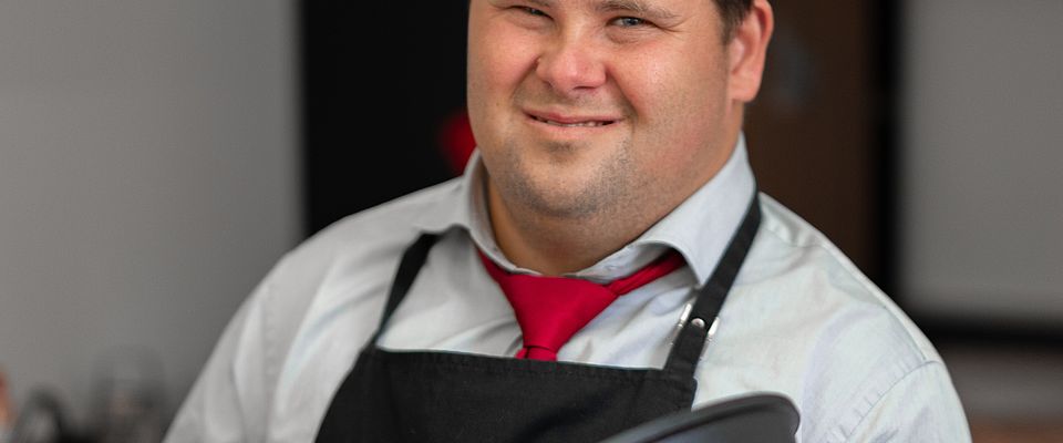 Junger Mann mit Down-Syndrom in hellblauem Hemd, roter Krawatte, schwarzer Schürze und schwarz-weißer Kochmütze hält mit beiden Händen eine schwarze Pfanne in der Hand und lächelt in die Kamera.