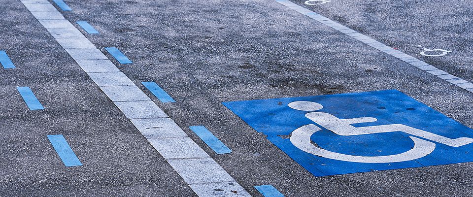 Ausschnitt einer asphaltierten Straße mit blauen Markierungen. In der Bildmitte sind zwei weiße Piktogramme auf blauem Untergrund zu sehen, die Behindertenparkplätze markieren.