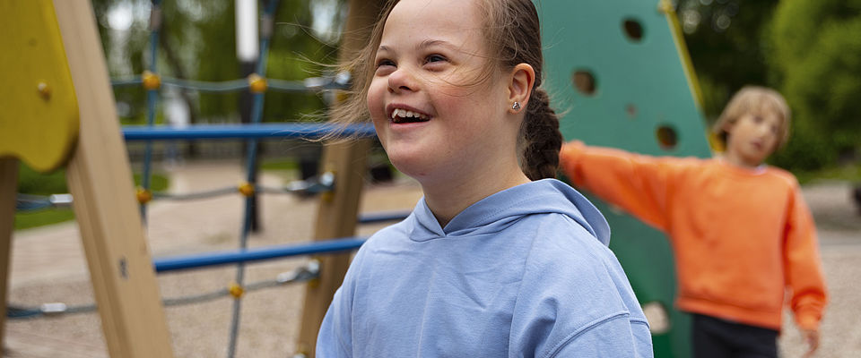 Lächelndes Mädchen mit Downsyndrom im Vordergrund auf einem Spielplatz. Es trägt einen hellblauen Kapuzenpullover und die dunkleblonden Haare zu einem Zopf gebunden. Im Hintergrund rechts hinter ihr ist ein blondes Kind in orangefarbenem Pullover zu sehen, das sich am Spielgerät abstützt.