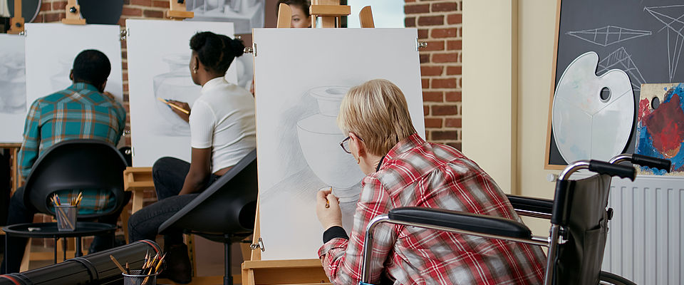 Blonde, kurzhaarige Frau im Rollstuhl nimmt an einem Zeichenkurs teil. Auf dem Bild ist die Frau in rotkariertem Hemd und schwarzer Hose damit beschäftigt, an der Staffelei eine Vase mit Bleistift zu zeichnen. Im Hintergrund sind verschwommen zwei weitere Kursteilnehmer in einem Lehrraum zu sehen.  