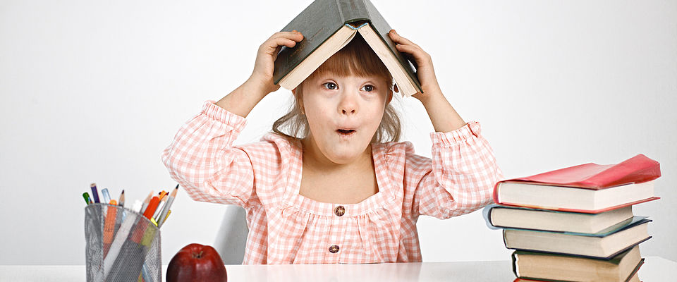 Ein Mädchen mit Downsyndrom in rosa-kariertem Kleid sitzt an einem weißen Tisch und hält sich ein aufgeklapptes Buch wei ein Dach über den Kopf. Ihre Lippen formt sie zu einem O. Links vor ihr steht ein Behältnis mit Stiften darin und ein roter Apfel, rechts vor ihr ist ein bunter Bücherstapel zu sehen.