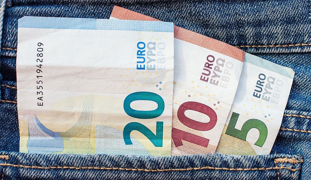In einer blauen Jeanshosentasche befinden sich drei Geldscheine: ein blauer 20-Euro-Schein, ein roter 10-Euro-Schein und ein grüner 5-Euro-Schein, die alle einander teilweise überlappend aus der Hosentasche herausschauen.