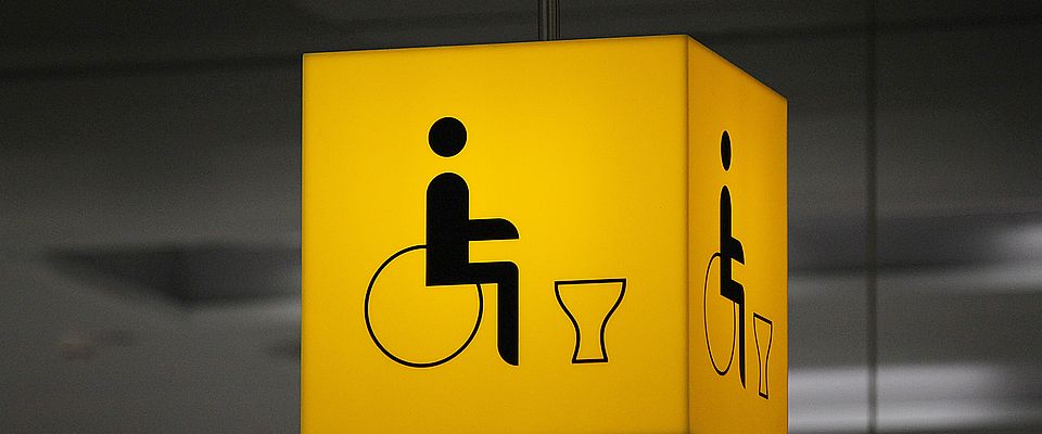 Gelbes, illuminierbares Element, abgehängt von der Decke. Auf dem gelben Grundkörper ist schwarz das Piktogramm für Behindertentoilette abgebildet.