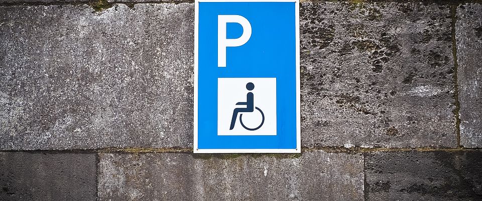 Blaues Schild für Behindertenparken, aufgebracht an einer Hauswand, die mit grauen Steinfliesen verkleidet ist. Auf dem Schild ist ein weißes P auf blauem Untergrund dargestellt, ferner ein weißes Quadrat, in dessen Mitte ein schwarzes Piktogramm eines Rollstuhlfahrers abgebildet ist. 