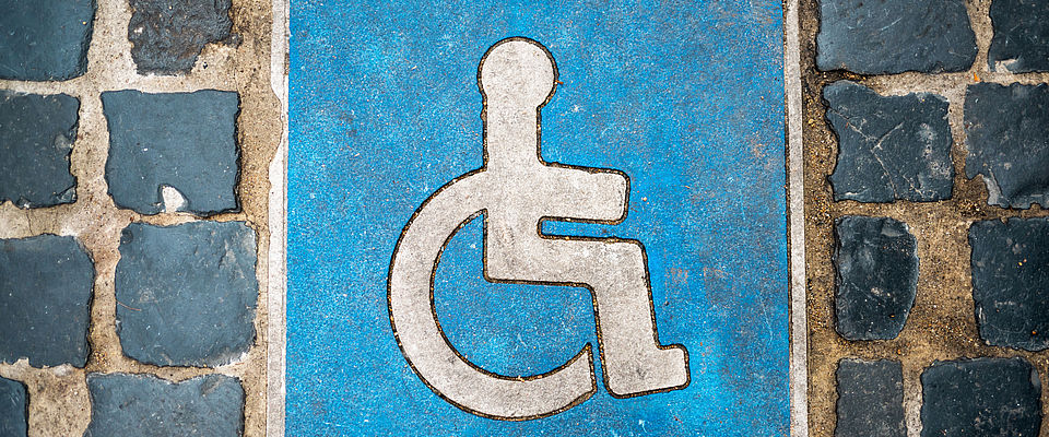 Weißes Piktogramm, das für Behindertenparkplatz steht, auf blauem Untergrund, eingefasst von kleinen schwarzen Pflastersteinen.